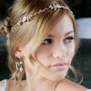 Bride looking away from the cameraposing with a crystal minimalist wedding hair wrap in her hair. Her hair is blonde and is up on a low soft messy bridal bun. In the background is a bed in the bridal suite.