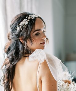 Bride wearing a bridal gown with large tulle bow at the shoulders. She has long curly dark hair and wears a Statement floral pearl bridal hair vine. Behind her are curtains to a large window.