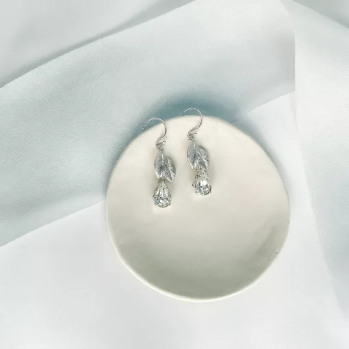 leaf and crystal drop bridesmaid earrings lying on a small clay round dish on top of a beige fabric background and light blue silk ribbon.