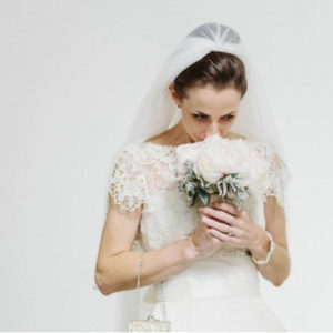 A bride, all in white with a veil, leans forward to smell her white flower bouquet