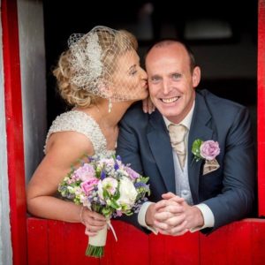 A bride stands sideways kissing a smiling groom leaning against a red stable door