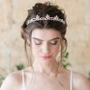 Bride looking down, wearing a strapped bridal gown. Her hair is up and she wears a Crystal Pearl handmade wedding tiara. In the background is a stone wall.
