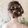 bride stood with her back to the camera. Her gown features straps. You can only see her shoulders. She faces a stone wall in a barn. Her hair is up in a curly messy updo with modern celestial starburst hair pins decorating it.