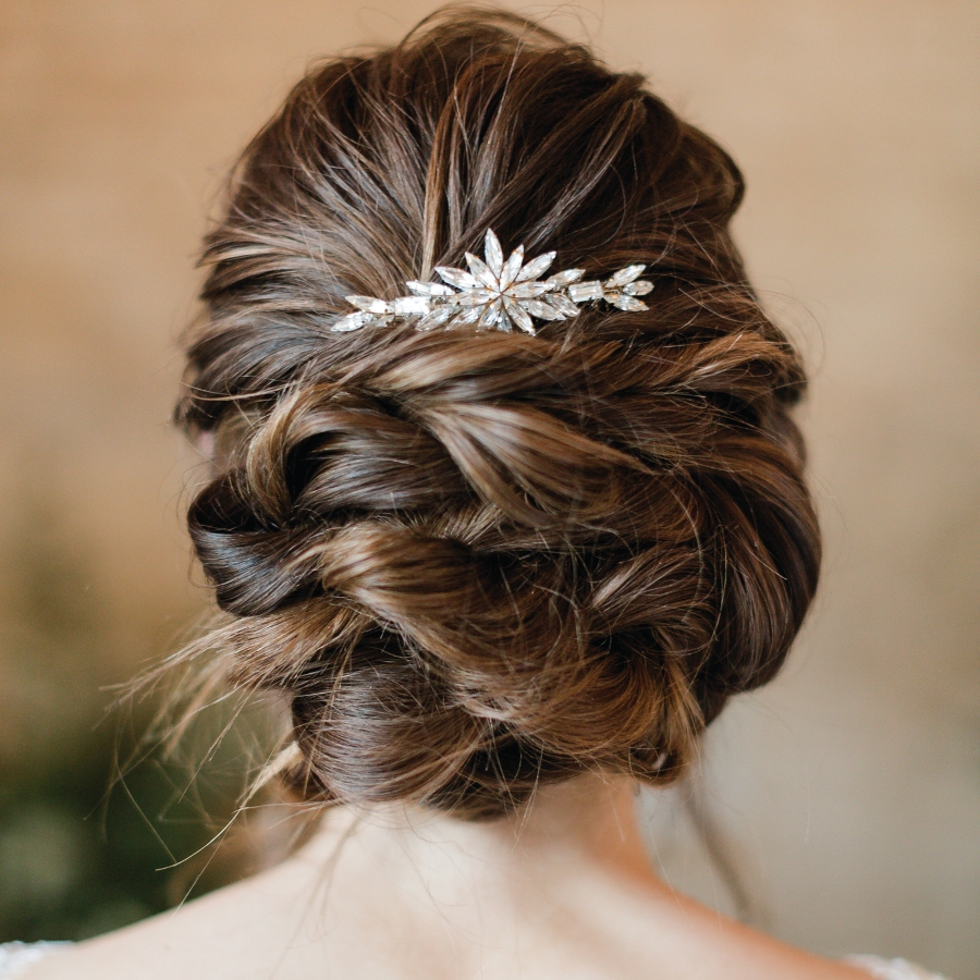 Shoulders and back of the head view of a bride. She stands in front of a stone wall. Her hair is dark and she has a curly bridal hair bun with Statement crystal wedding hair comb in.