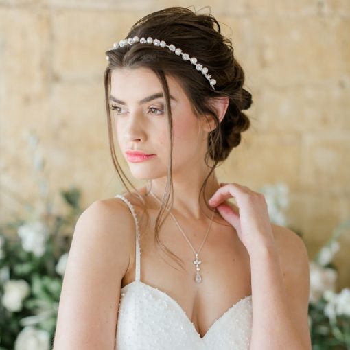 Woman stood with ivory wedding dress with straps, wearing a simple pearl wedding alice band with dark brown hair in a low braided updo.