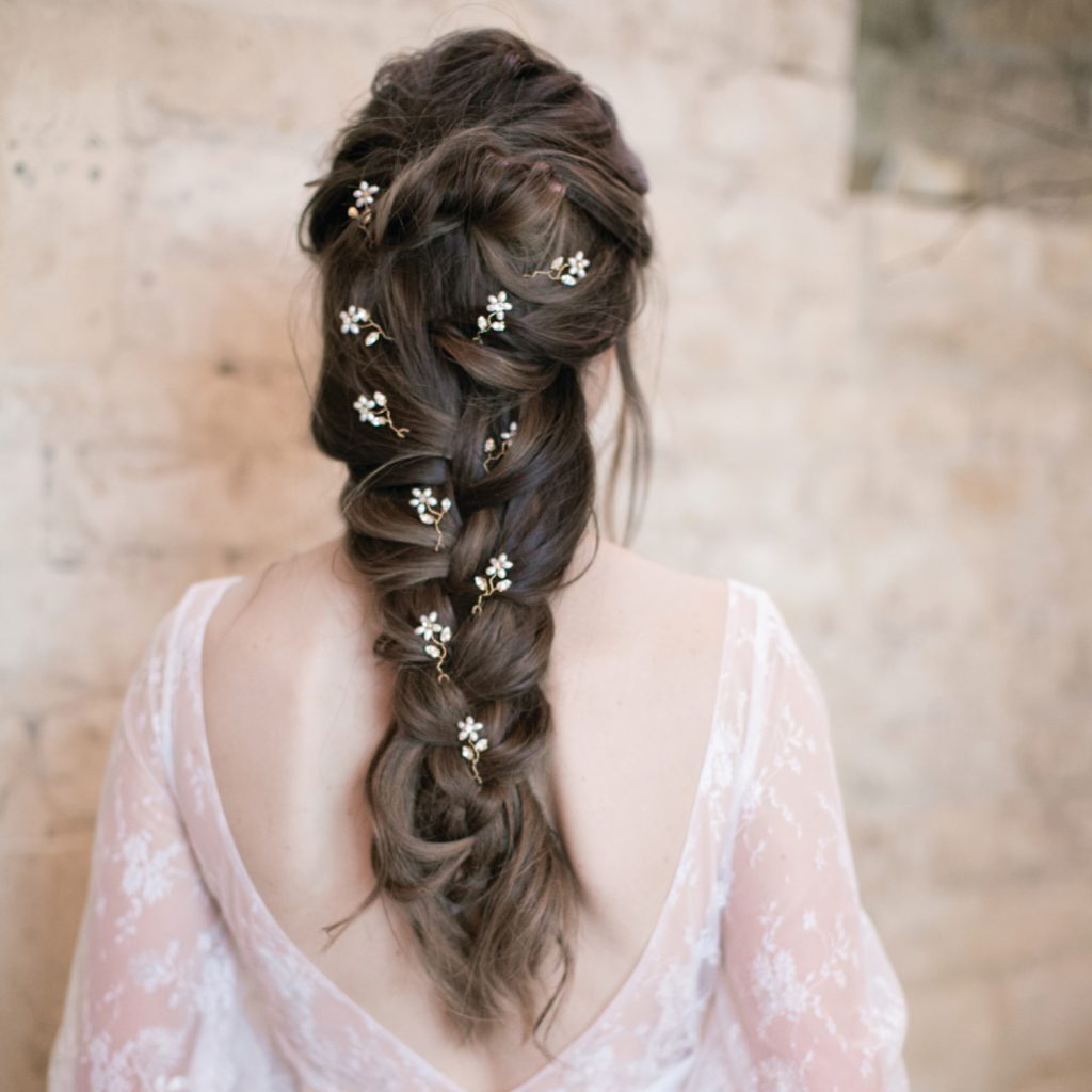 Stone wall in background. Woman standing with her back to the camera in a low back, lace wedding gown and long loose braided hair with Small dainty blossom bridal hair pins scattered through.