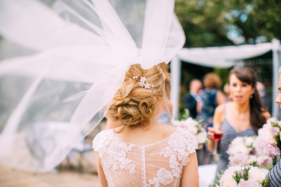 Birde stood with her back to the camera. Her veil has been caught by the wind and lifts high abover her showing her undone hair up bridal hair do, with a floral hair pin.