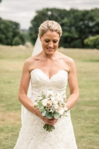 Bride stood in front of the camera looking down at her modest bridal bouquet of whites and foliage, with a lawn and trees behind