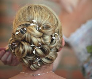 Bride having makeup applied . Her back is to the camera. You can see a full and curly chignon of blonde hair with lots of leaf and diamante hair pins cattered through.