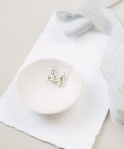 sterling silver Swarovski crystal earrings set on a small cream coloured bowl, on a white paper wedding invitation with blue ribbon. The background is a beige fabric.