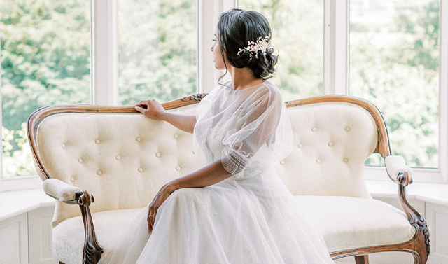 Wedding Hair Accessories and Jewellery. Image of woman in a bridal gown sat on a cream upholstered day sofa looking out of a grand window at the garden outside.