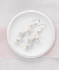 Small cream dish with bridal flower and pearl drop earrings laying on top. Underneath the dish is a beige fabric and blush pink silk ribbon