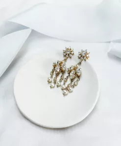 Image shows white fabric background with blue silk ribbon draped across, with small clay bowl on top with Victorian style crystal and pearl chandelier earrings