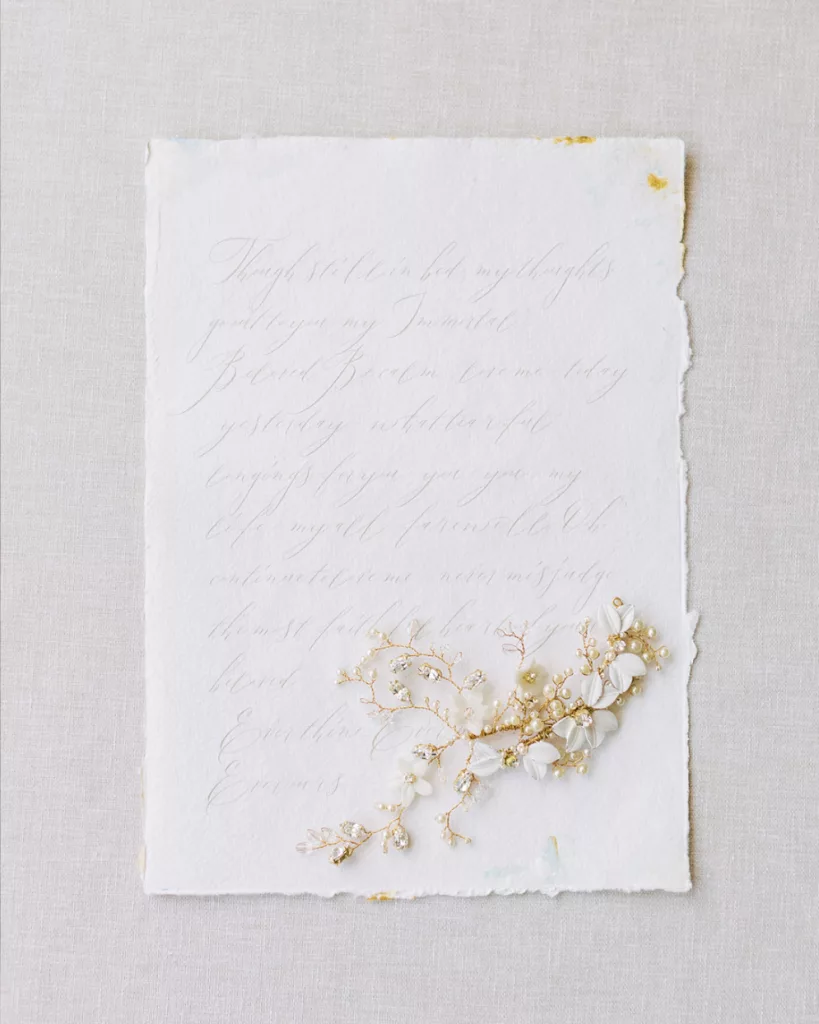 letter on flat surface written with calligraphy with slightly damaged edges to look old. Placed on top are two bridal hair accessories. Both hair vines with a mix of pearls, enamelled white leaves and diamante