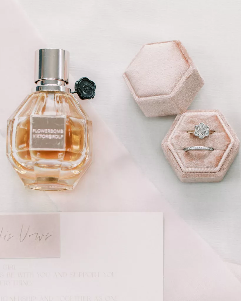 Viktor & rolf perfume bottle next to a blush pink ring box with platinum wedding and engagement ring and a blush pink and white invitation below on a flat surface