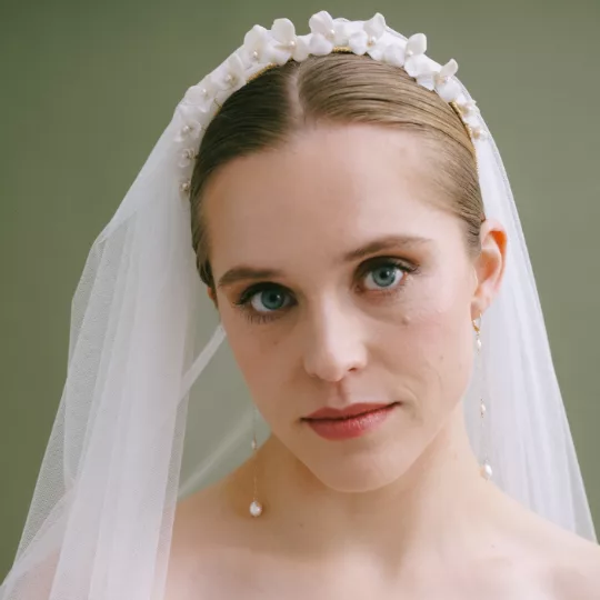 Bride wearing a tiara with flowers and veil. She looks at the camera and wears long dangly pearl earrings