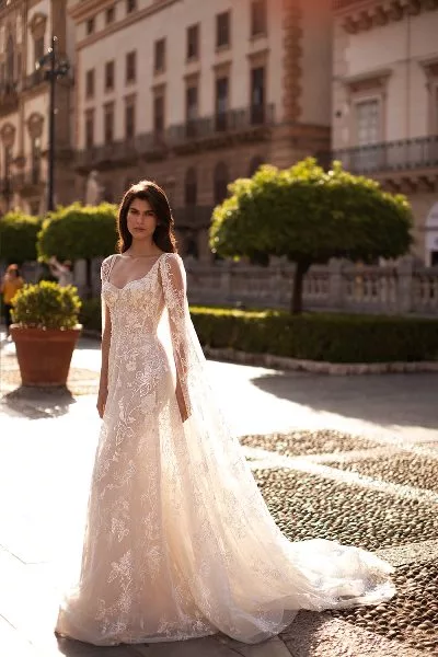 Bride standing in a courtyard,, with trees and buildings behind. She wears a white bridal gown, with cape.