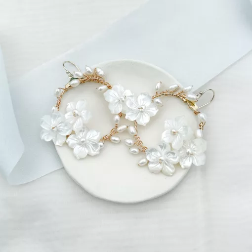 Large delicate handmade floral pearl hoop wedding earrings set on a cream dish, overlaying an ivory fabric background with light blue silk ribbon.