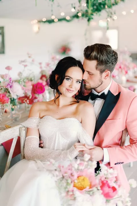 dark haired woman in white bridal gown sat at wedding breakfast table with dark blonde hair groom wearing a velvet pink tuxedo jacket and white shirt. The background features lots of pink flowers in various shades.