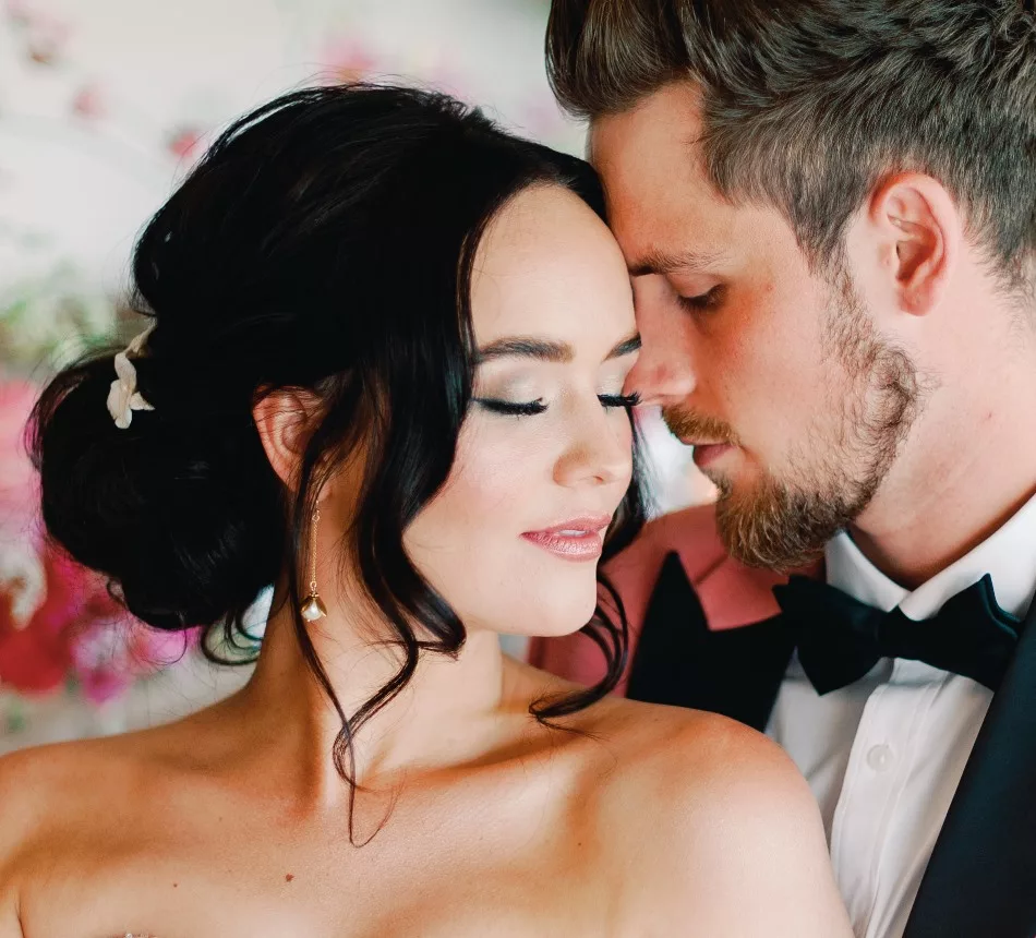Dark haired bride in loving pose with groom. Their heads are touching, and eyes are closed. The groom wears a black bow tie, and velvet pink suit jacket.