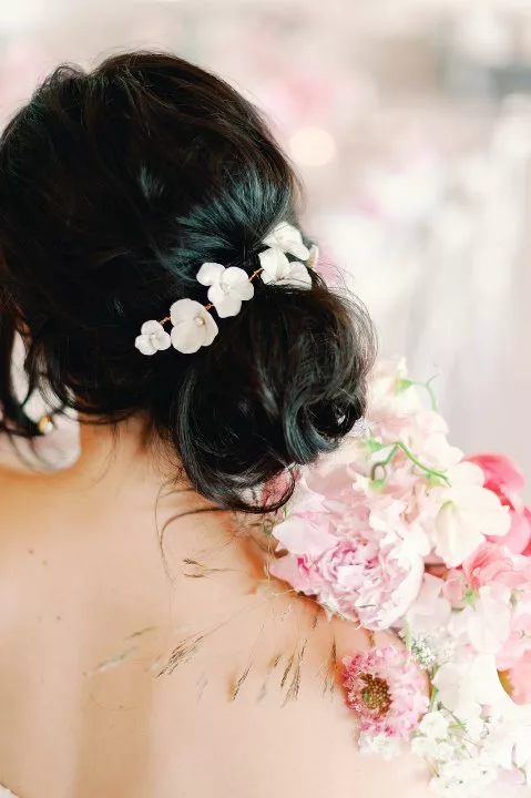Woman with dark hair in a low, messy bun. The back of her head is decorated with a large flower hair vine, She holds a pink flower bouquet over one shoulder.