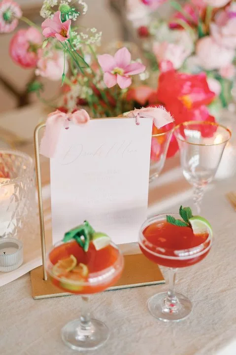 Wedding breakfast table set up. 2 cocktail glasses with a pink punch drink, and decorated with citrus fruits stand in front of a drinks menu. in the background are lots of pink flowers.