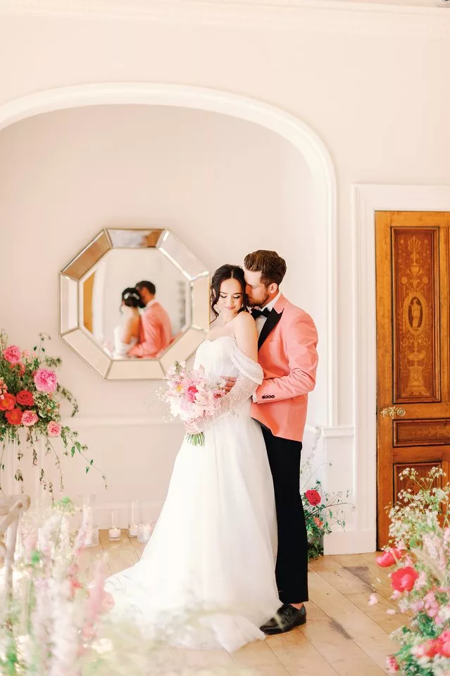 Bride and groom (in pink suit) standing in a ceremony space surrounded by an abundance of large pink flowers on display, next to the guest seating.