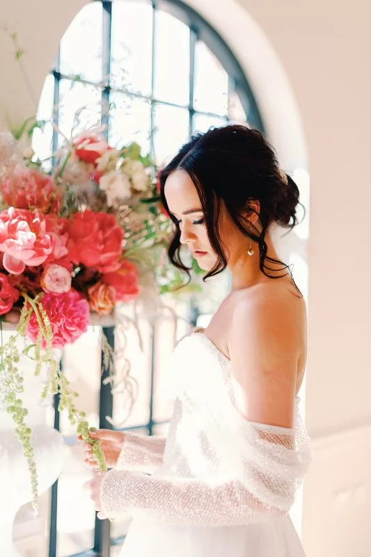 Bride looking at a tall floral vase display of pink flowers, behind is an arched window.