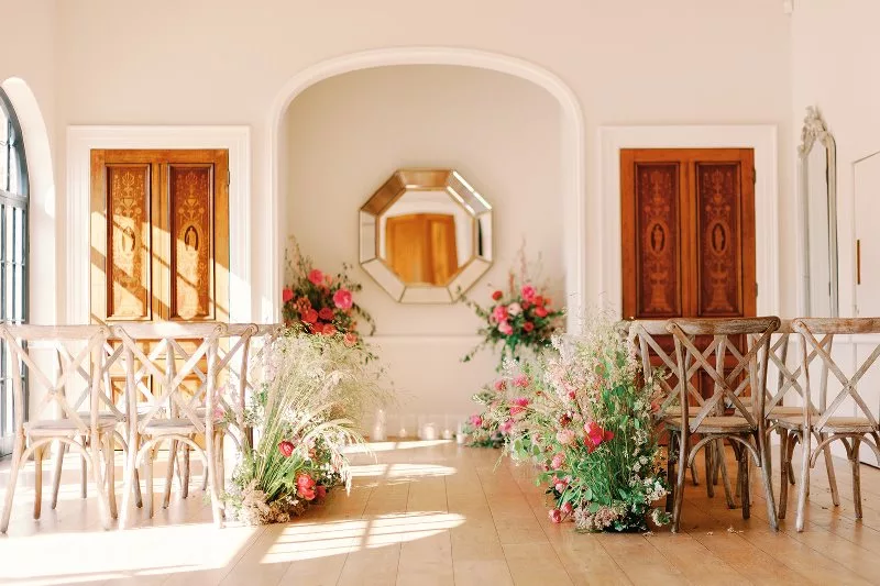 Ceremony room filled with light. A ceremony space in the middle is flanked either side with large floral pink displays and 2 wooden doors. Each side has sets of guest seating with a meadow of flowers parallel.