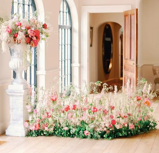 Orangery room with arched windows to the left. A tall white urn sits in front with a large bouquet of flowers. On the floor in the centre lies an abundance of pink flowers in a flower meadow.