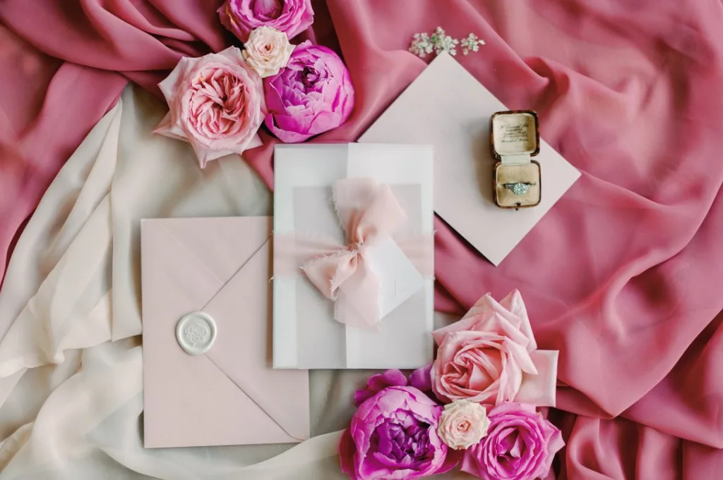 Flatlay image showing different shades of pink materials, and a mix of wedding stationery placed over the top, with pink wedding flowers in clusters. A small ring and old ring box sit on one of the wedding invites.