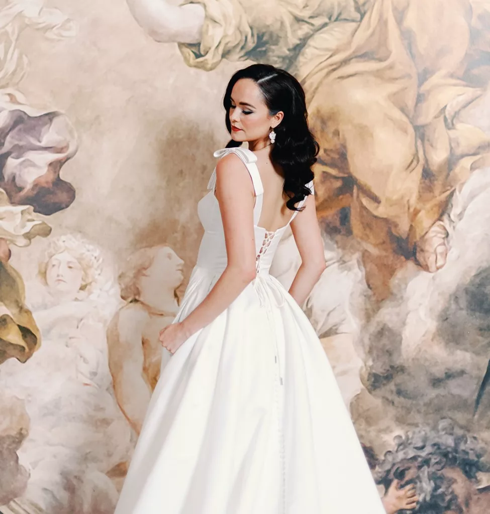 Dark haired woman with hollywood long hair curls, She wears a wedding dress in ivory, with large straps and bows. She stands in front of a large oil painting of flowers.