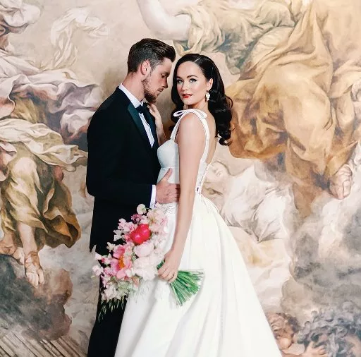 Dark haired woman with hollywood long hair curls, She wears a wedding dress in ivory, with large straps and bows. She stands in front of a large oil painting of flowers embracing a groom with a black dinner suit.
