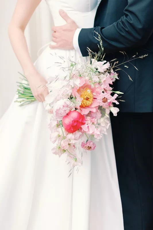 flower bouquet of pink peonies, roses and sweetpea flowers with a bride and groom standing behind.
