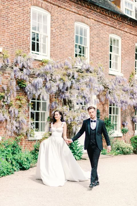Bridal and Groom walking in front of a large manor house, with wisteria trailing the windows.