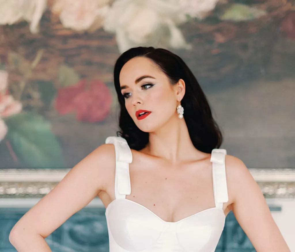 Dark haired woamn with hollywood long hair curls, She wears a wedding dress in ivory, with large straps and bows. She stands in front of a large oil painting of flowers.