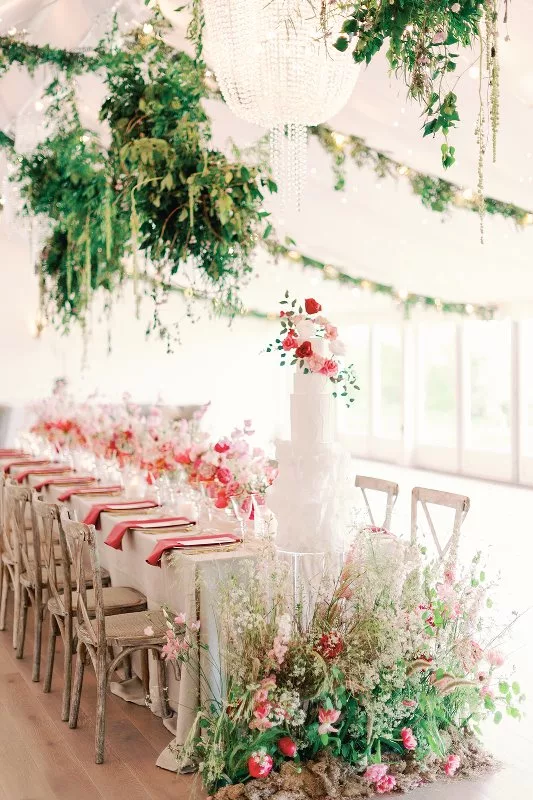 Inside a marquee, a long table decorated for a wedding breakfast, with pink flowers, and lots of green foliage hangs from the ceiling. A 3 tier white wedding cake with pink flowers sits at the end with a meadow of pink flowers on the floor in front of it.