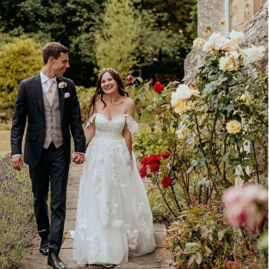 Bride and groom walking in the grounds of a garden. To their right are a collection of different colour rose bushes, and to the left of the path are bushes of lavender.