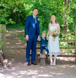 Bride and groom with little page boy in blue standing in front of a wooden gate in woodland.