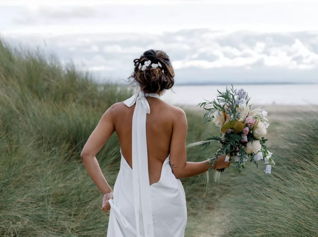 Wedding accessories inspired by nature - Woman in wedding dress walking across the grassy sandunes down to a beach, holding a wedding bouquet.