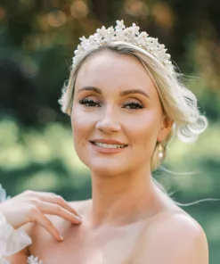 Woman with blonde hair and lace bridal dress smiling and wearing a Flower pearl bridal crown, and pearl earrings. Grounds of a country house garden are in the background.