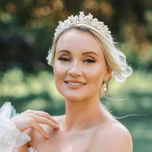 Woman with blonde hair and lace bridal dress smiling and wearing a Flower pearl bridal crown, and pearl earrings. Grounds of a country house garden are in the background.