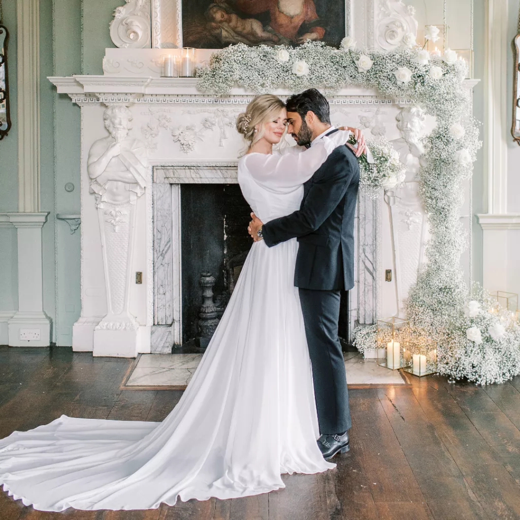 bride and groom stood in front of a large georgian fireplace that is decorated with Shades of green wedding gypsophila and white roses with lit candles.