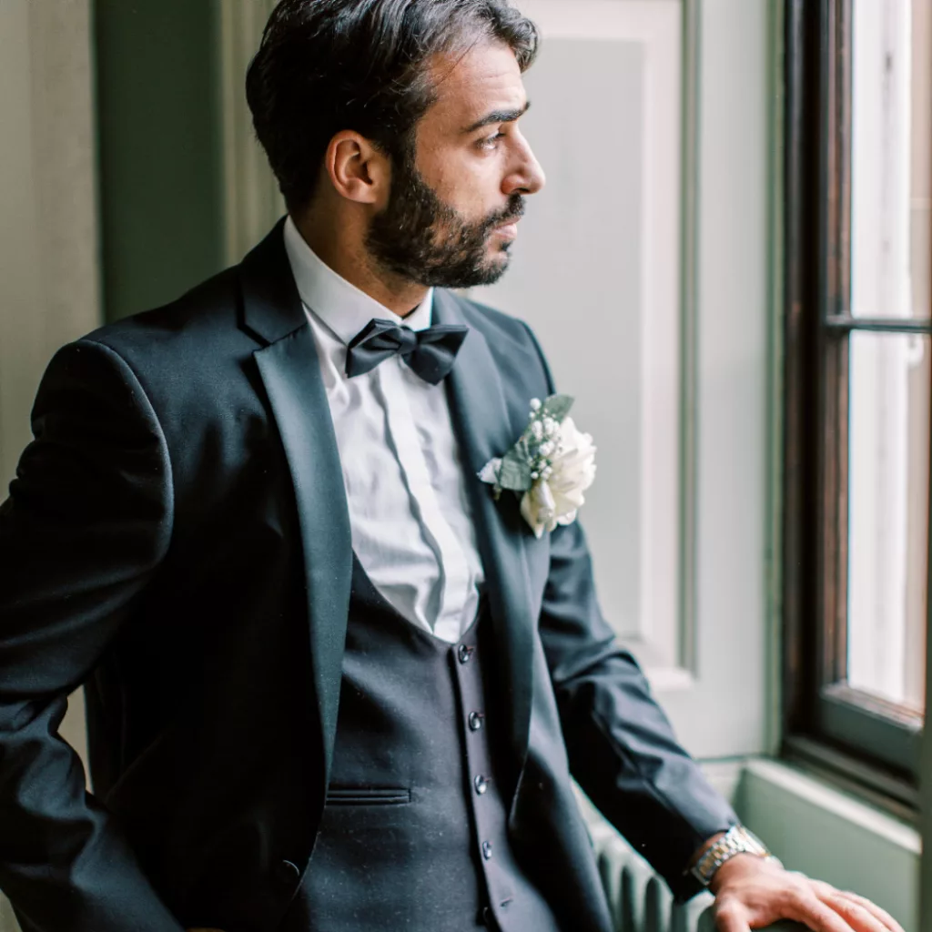 Groom with dark hair resting his hand on a green radiator looking out of the window. He wears a black suit, waistcoat and black bow tie, and white rose button hole.