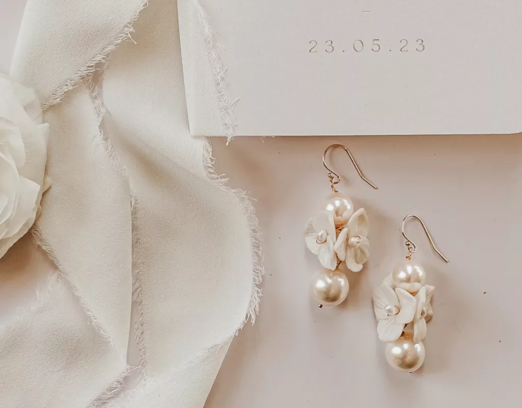 A guide on types of earring fittings with image of a nude background with floral earrings with pearls, decorated with ivory silk ribbon and a section of a wedding invitation.