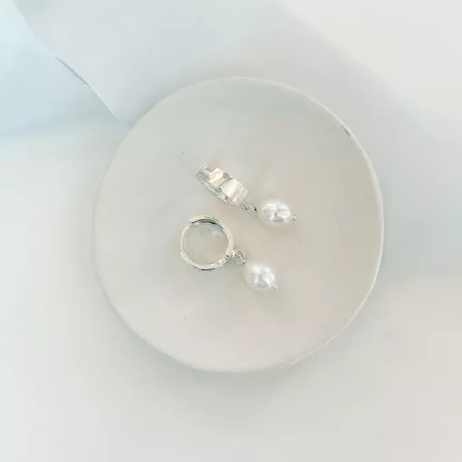 white background with light blue ribbon draped over it. A cream clay dish sits on top with a pair of Huggie pearl drop wedding earrings in silver