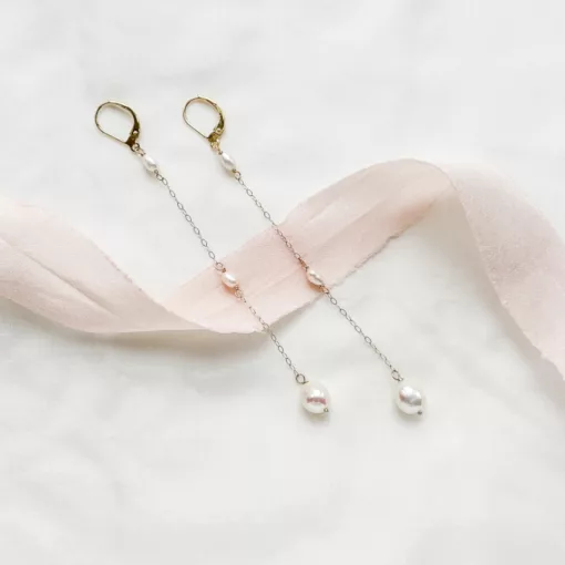 long drop pearl bridal earrings lying on a soft ivory material with blush ribbon draped across