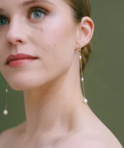 Long drop pearl bridal earrings being worn by a model with her hair in a bun, against a green background