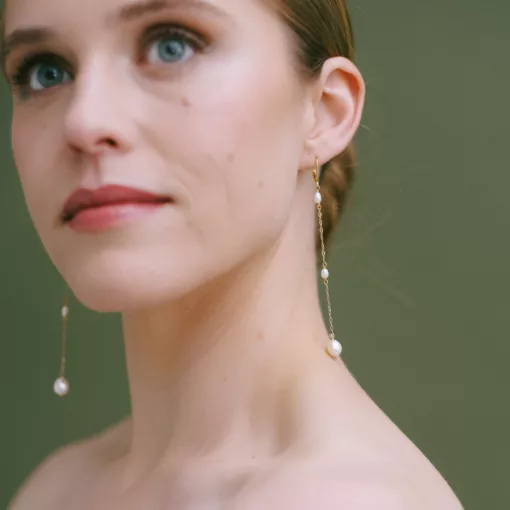 Long drop pearl bridal earrings being worn by a model with her hair in a bun, against a green background