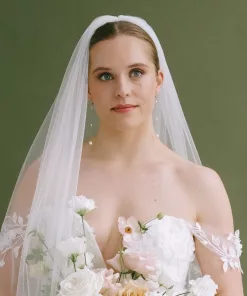 Long drop pearl bridal earrings being worn by a bridal model, wearing a veil and has her hair back in a bun. She wears a bridal gown with a straps that drape below her shoulders and she holds a bridal bouquet with peach and white flowers
